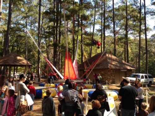 A lively outdoor scene with people gathered around a bungee trampoline attraction in a wooded area.