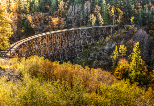 A wooden trestle bridge arches over a colorful autumn landscape with trees in shades of orange and green.