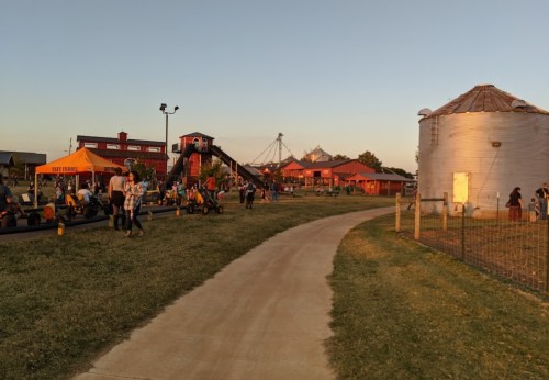 A pathway leads through a farm amusement area with people, rides, and a silos at sunset.