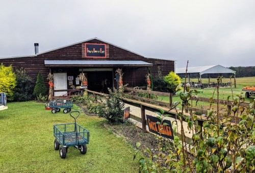 A rustic barn with a sign, surrounded by greenery and a cart, set in a rural landscape under a cloudy sky.