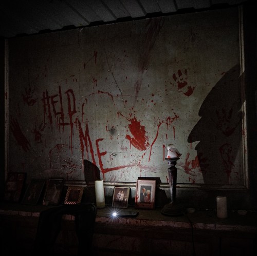 A dimly lit room with a wall stained in red, featuring handprints and the words "HELP ME," alongside framed photos and candles.