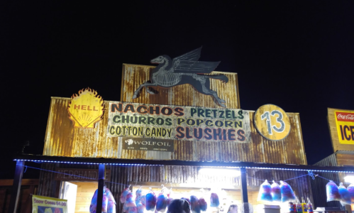 A brightly lit food stand at night featuring signs for nachos, pretzels, churros, popcorn, cotton candy, and slushies.