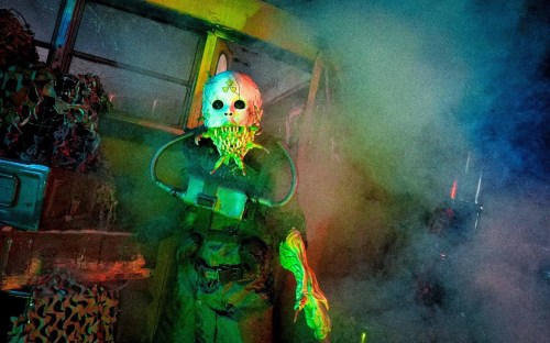 A creepy figure in a gas mask and skull mask stands in a smoky, colorful environment, evoking a horror theme.