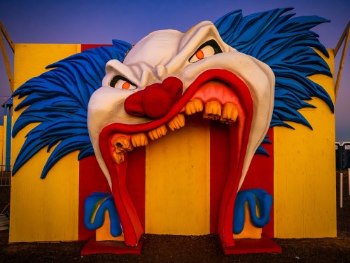 A large, colorful clown face sculpture with an open mouth, set against a striped background at dusk.