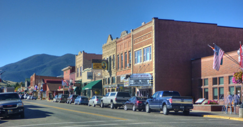 A charming small-town street lined with shops, parked cars, and mountains in the background under a clear blue sky.