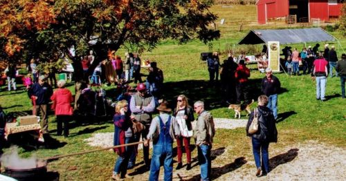 A lively outdoor market scene with people socializing, a red barn in the background, and a dog nearby.