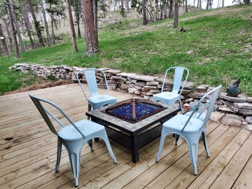 A cozy outdoor seating area with four blue chairs around a fire pit on a wooden deck, surrounded by trees.