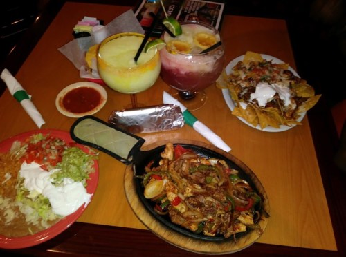 A table spread with sizzling fajitas, nachos, a salad, a burrito, and colorful cocktails.