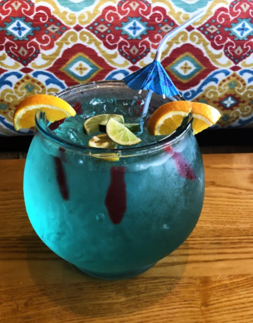 A colorful cocktail in a fishbowl glass, garnished with orange and lime slices, and a small blue umbrella.