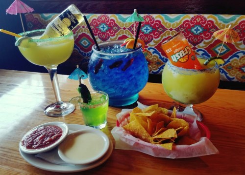 A colorful assortment of cocktails with umbrellas, a shot, and a basket of nachos with salsa and cheese dip.