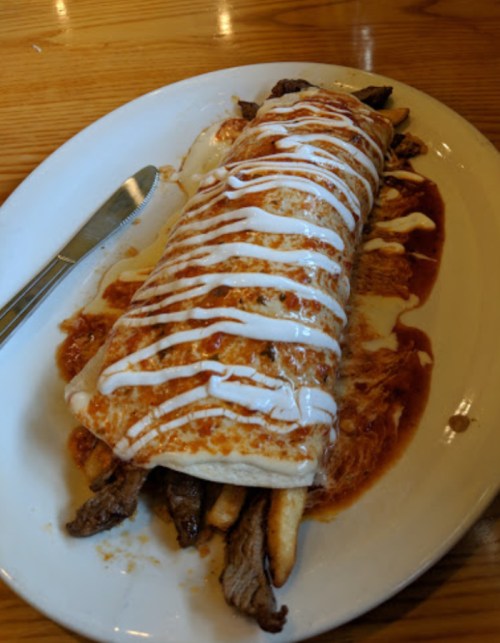 A large burrito filled with meat, topped with sauce and drizzled with white cream, served on a plate with fries.