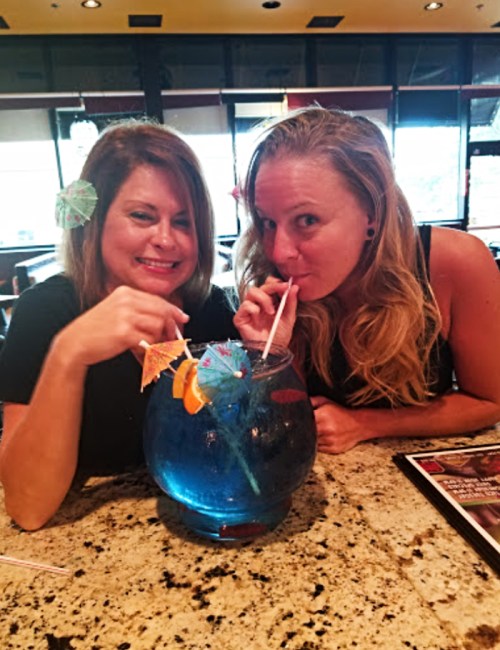 Two women smiling and sipping from a large blue drink with colorful garnishes at a restaurant.
