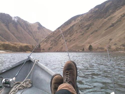 A pair of boots resting on a boat, with fishing rods and a scenic view of mountains and water in the background.