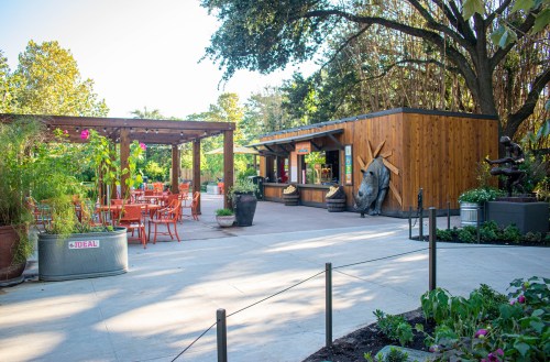 A sunny outdoor area featuring a wooden building, seating, and lush greenery, creating a welcoming atmosphere.
