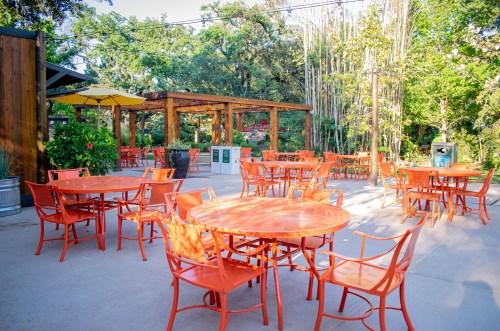 Outdoor seating area with orange tables and chairs surrounded by greenery and wooden structures.