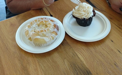 A cinnamon roll and a chocolate cupcake with frosting on white plates, placed on a wooden table.