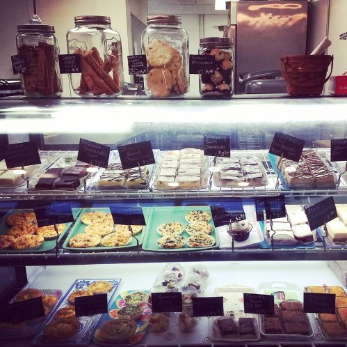A display case filled with various baked goods, including cookies, bars, and pastries, labeled with small chalkboard signs.