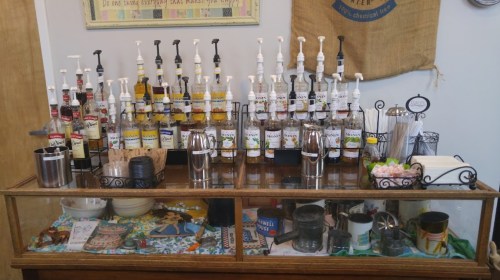 A display of various syrup dispensers and coffee accessories on a wooden counter in a café setting.