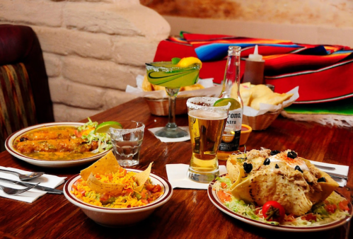 A table set with Mexican dishes, including tacos, enchiladas, a drink, and a dessert, surrounded by colorful decor.