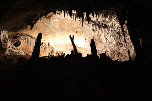 Silhouette of stalagmites and stalactites in a dimly lit cave, with a bright area illuminating the cave's ceiling.