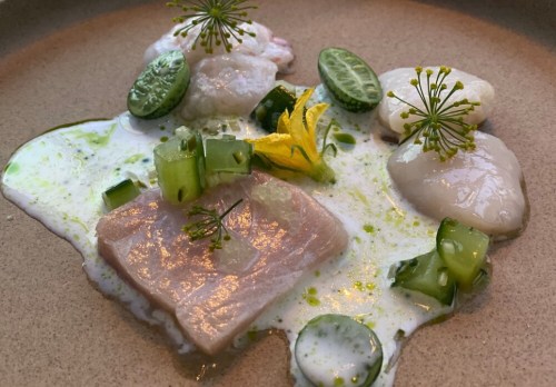 A gourmet dish featuring fish, green vegetables, and edible flowers on a creamy sauce, artfully arranged on a plate.