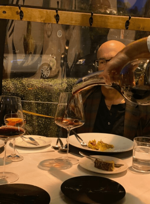 A hand pours red wine into a glass at a dining table with plates and cutlery, while a person sits in the background.