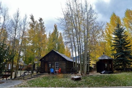 A rustic log cabin surrounded by colorful autumn trees and a small gazebo in a serene outdoor setting.