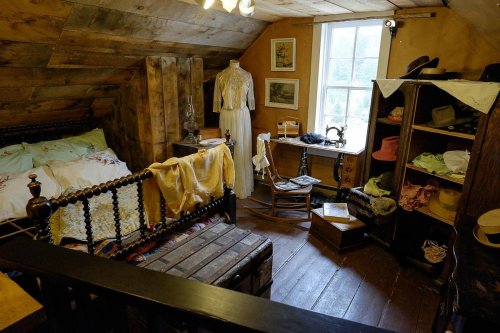 A cozy attic room with a bed, vintage clothing, a sewing machine, and various hats displayed on shelves.