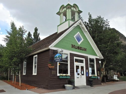A charming museum building with green and brown siding, featuring a bell tower and surrounded by trees.