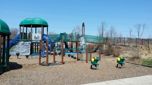 A colorful playground with slides, climbing structures, and spring riders, surrounded by grass and trees on a sunny day.