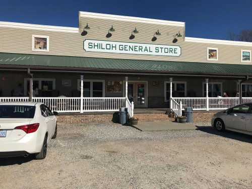 Exterior of Shiloh General Store with a green roof, white porch, and parked cars in front on a sunny day.