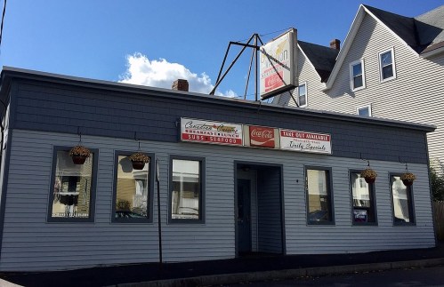 A gray building with a sign for a sandwich shop, featuring large windows and hanging flower baskets.