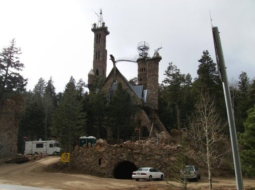 A unique stone castle with towers, surrounded by trees, and a vehicle parked nearby on a dirt road.