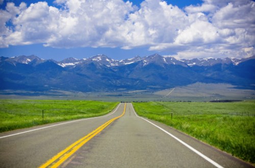 A long, empty road stretches towards majestic mountains under a blue sky with fluffy clouds. Lush green fields flank the road.