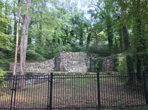 Stone ruins surrounded by lush greenery, with a black fence in the foreground.