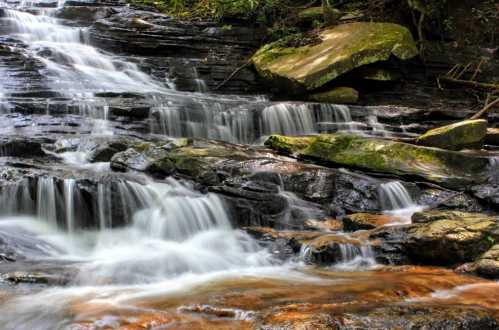 A serene waterfall cascades over smooth rocks, surrounded by lush greenery and a hint of orange in the water.