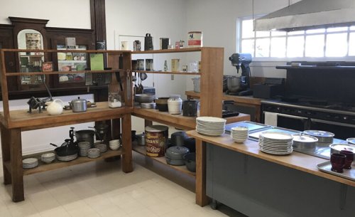 A spacious kitchen with wooden shelves, various cookware, and a large stove, featuring organized plates and utensils.