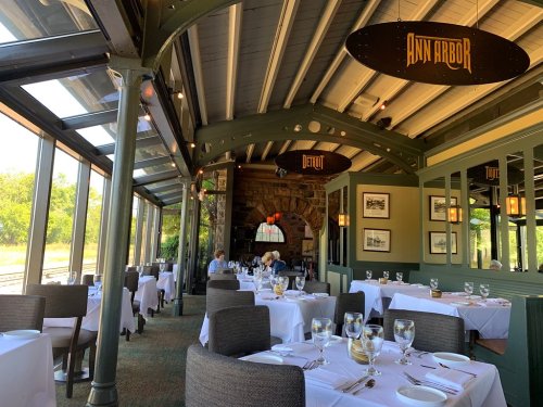 A bright restaurant interior with tables set for dining, featuring large windows and decorative signs for Ann Arbor and Detroit.