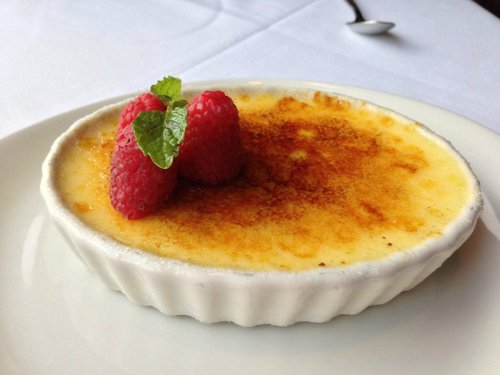 A creamy dessert topped with caramelized sugar, garnished with raspberries and a mint leaf, served in a white dish.