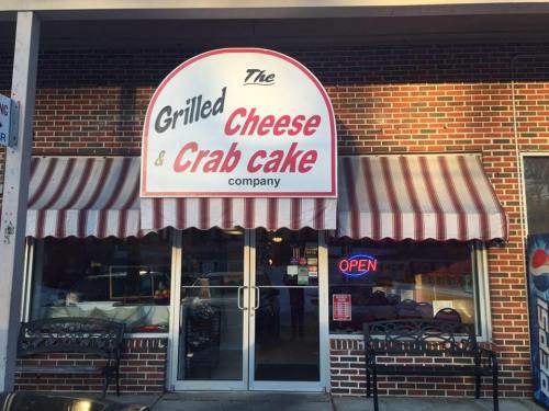 Exterior of a restaurant with a sign reading "The Grilled Cheese & Crab Cake Company" and an "OPEN" neon sign.
