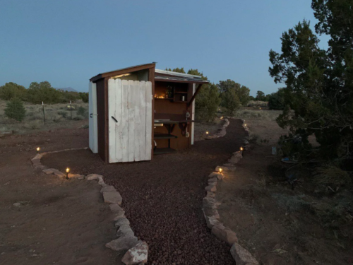 A small wooden shed with a white door, surrounded by a gravel path and lit by soft lights, set in a natural landscape.