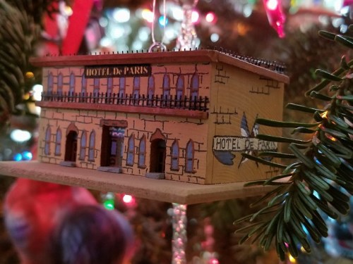 A decorative ornament of the Hotel de Paris, featuring a detailed building design, hanging on a Christmas tree.