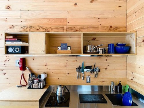 A wooden kitchen with open shelves displaying cookware, books, and a vintage phone against a wood-paneled wall.