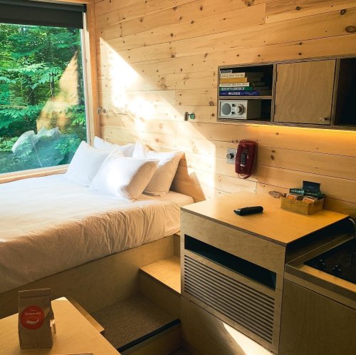 Cozy wooden cabin interior with a bed, shelves, and a large window overlooking greenery.