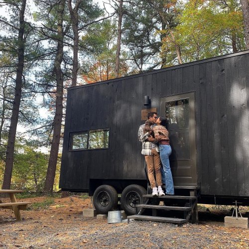 A couple embraces on the steps of a tiny house in a wooded area, surrounded by trees and outdoor seating.