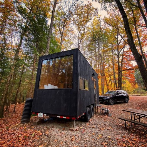 A black tiny house on wheels surrounded by autumn trees, with a car parked nearby and picnic tables in the foreground.