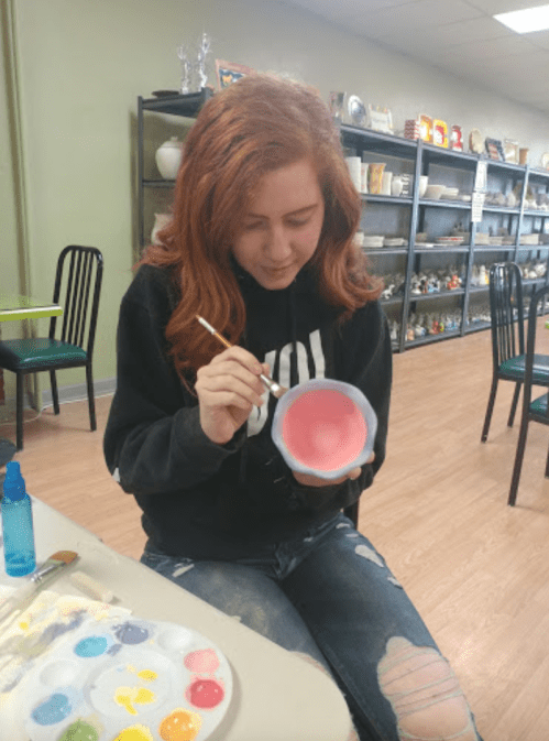 A person with red hair paints a bowl in a pottery studio, surrounded by colorful paints and pottery items.