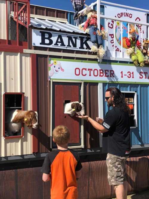 A child interacts with a man at a petting zoo, feeding goats through a bank-themed structure. Fall decorations are visible.