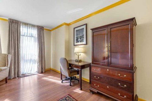 A well-lit room featuring a wooden desk, chair, dresser, and large window with sheer curtains.