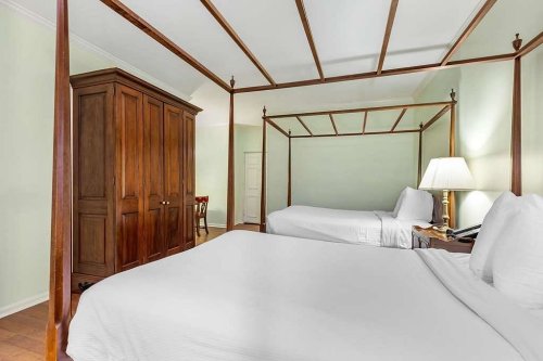 A spacious hotel room featuring two canopy beds, a wooden wardrobe, and a bedside lamp against a light green wall.